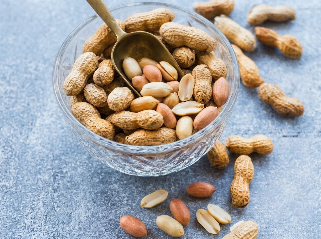 Groundnut kernels and whole peanuts in a glass bowl with scattered peanuts on a blue surface.